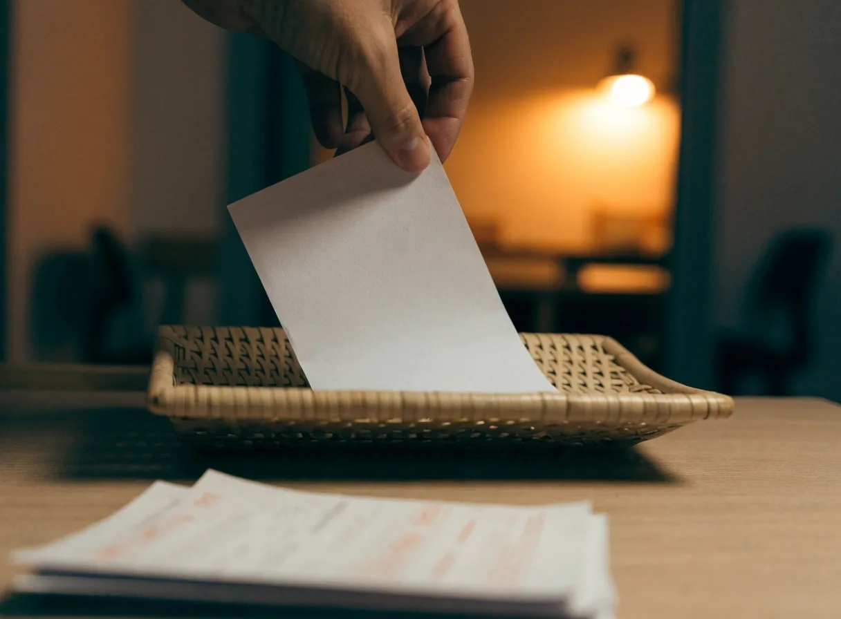 A paper invoice held between two fingers above a desk tray with receipts blurred in front